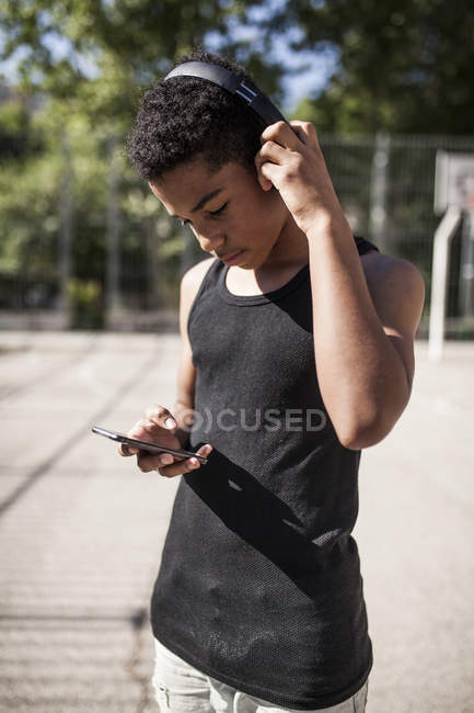 Afro young boy listening to music with smartphone and headphones on basketball court — Stock Photo