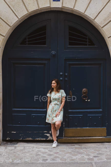 Woman in patterned summer dress with floral print leaning on shabby door and looking away — Stock Photo