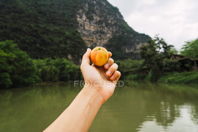 Mano umana che tiene succosa pesca su sfondo sfocato della superficie dell'acqua del fiume Quy Son, montagna e alberi, Guangxi, Cina — Foto stock