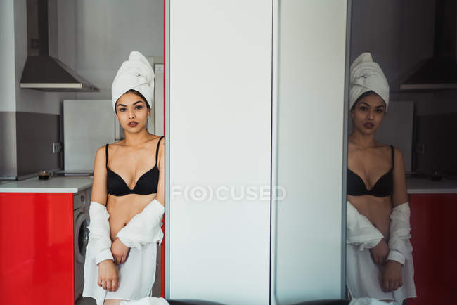 Sensual mujer joven en lencería y toalla en la cabeza apoyada en la pared en casa - foto de stock