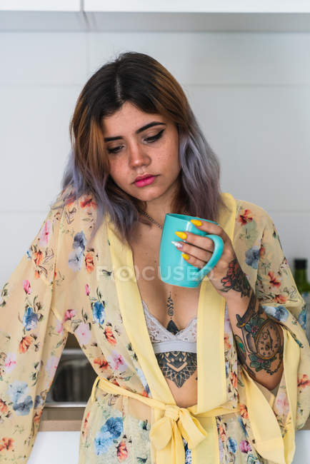 Mujer joven y somnolienta en bata de seda sosteniendo taza de bebida caliente mientras está de pie en la cocina por la mañana - foto de stock
