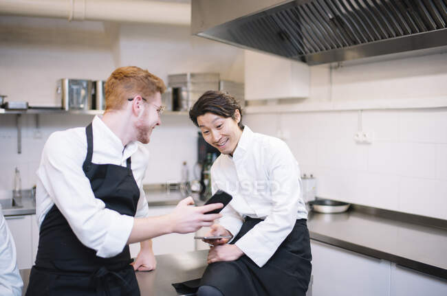 From below shot of two guys in cook uniform standing on restaurant kitchen and browsing smartphones during break — Stock Photo