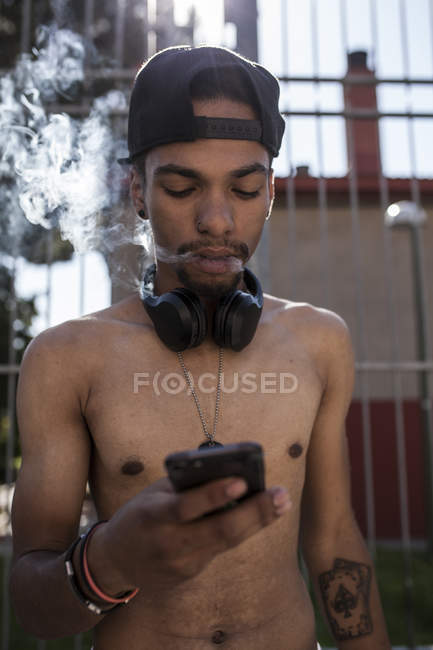 Afro jeune garçon écouter de la musique avec smartphone et écouteurs tout en fumant devant la grille — Photo de stock