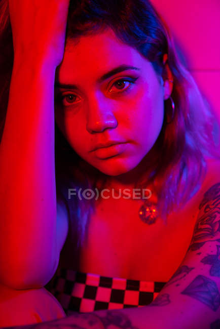 Jovem mulher tocando cabeça e olhando para a câmera enquanto sentado no quarto iluminado com luz vermelha — Fotografia de Stock