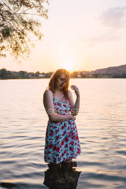 Mujer sensual en vestido de verano de pie en el agua del lago al atardecer - foto de stock