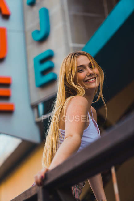 Smiling young woman leaning on fence against building and looking at camera — Stock Photo