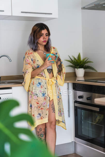 Татуированная вдумчивая молодая женщина стоит на кухне и расслабляется с чашкой горячего напитка — стоковое фото