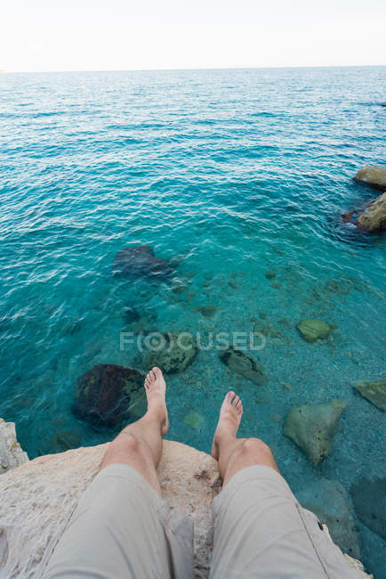 Ноги людини, що сидить на скелі над бірюзовою водою — стокове фото
