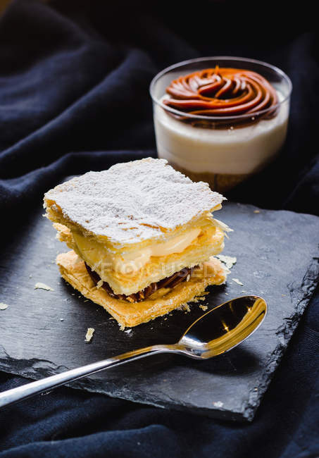 Primo piano del dolce sfoglia con panna su ardesia e dessert in tazza su tessuto nero — Foto stock