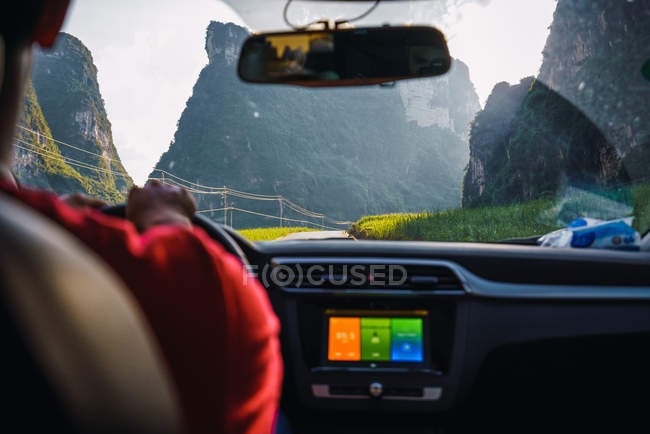 Primer plano del hombre que conduce el coche en la carretera estrecha entre las altas montañas impresionantes y los campos de hierba en el día soleado, Guangxi, China - foto de stock