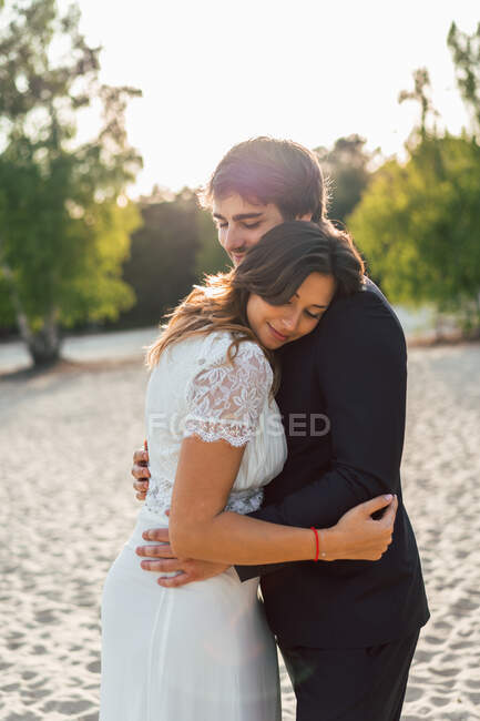 Amare l'uomo abbracciare bella sposa in abito elegante e guardarsi mentre in piedi sulla costa sabbiosa alla luce del sole — Foto stock