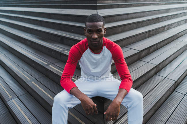 Sporty ethnic man sitting on outdoors stairs and looking at camera — Stock Photo