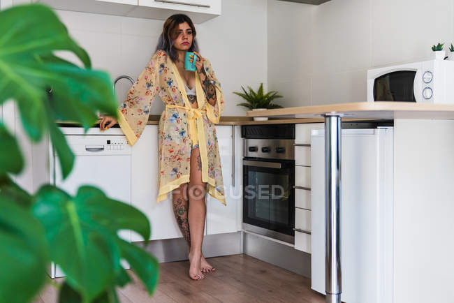 Tatuado mujer joven reflexiva de pie en la cocina y relajarse con una taza de bebida caliente - foto de stock