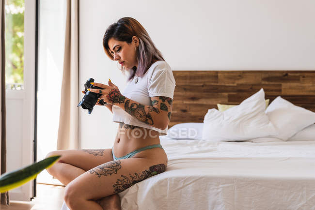 Mujer joven en bragas y camiseta mirando imágenes en la pantalla de la cámara profesional en la cama - foto de stock