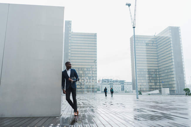 Афроамериканский бизнесмен опирается на стену на открытом воздухе с современными зданиями на заднем плане — стоковое фото