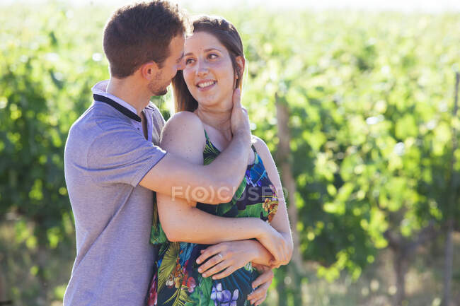 Vista laterale di felice giovane coppia con bicchieri di vino abbracciando e accarezzando teneramente mentre in piedi sul paesaggio con vigneto — Foto stock
