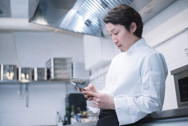 Вид сбоку красивый азиат в униформе шеф-повара, стоящий на кухне ресторана и просматривающий смартфон — стоковое фото