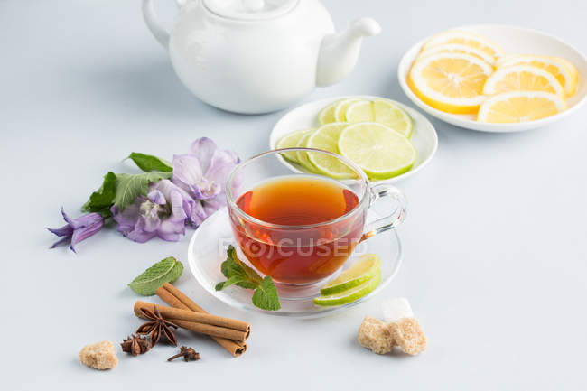 Schwarzer Tee in Glasschale auf Untertasse auf weißem Hintergrund mit braunem Zucker, Zimt, Minze, Blumen und Zitrusfrüchten mit Teekanne — Stockfoto