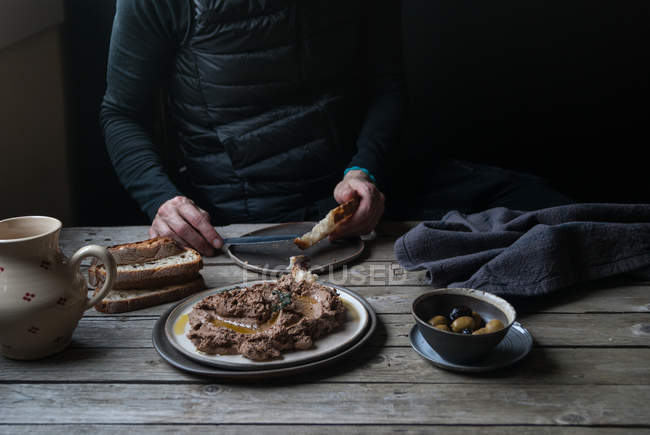 Чоловічі руки розкладають сочевичний паштет на хлібі на сільському дерев'яному столі — стокове фото
