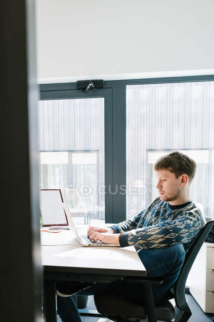 Seitenansicht eines Mannes in lässiger Kleidung, der bequem auf einem Bürostuhl sitzt und auf der Laptop-Tastatur auf einem weißen Schreibtisch tippt, mit großem Fenster im Hintergrund, aufgenommen durch offene Tür — Stockfoto
