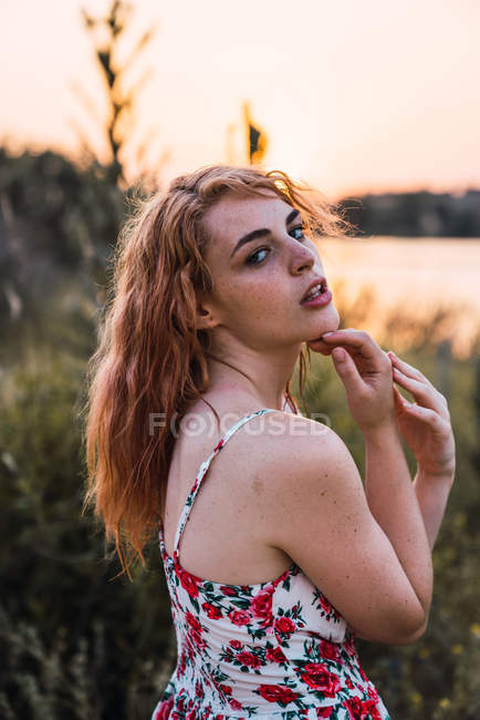 Jovem mulher sensual com sardas no vestido tocando o rosto de forma fascinante e olhando para a câmera ao pôr do sol — Fotografia de Stock