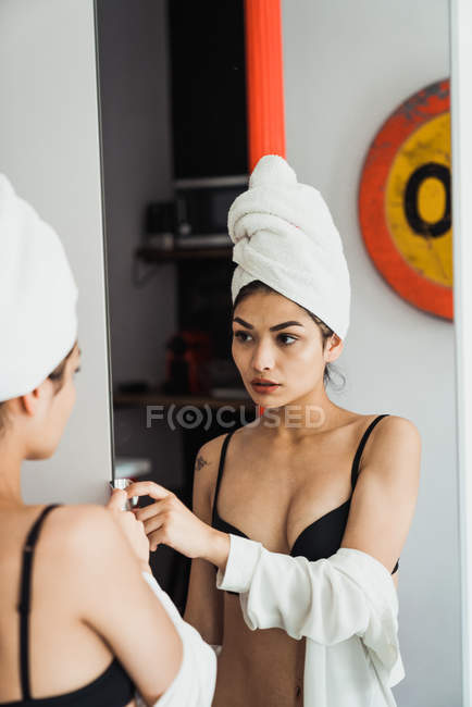 Jovem mulher em roupa interior preta e toalha na cabeça de pé no banheiro e olhando no espelho — Fotografia de Stock