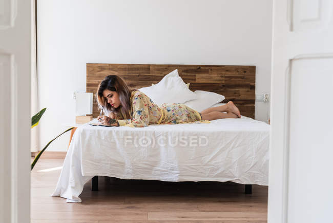 Молода жінка в шовковому халаті лежить на ліжку і робить ескізи в блокноті в стильній спальні — стокове фото