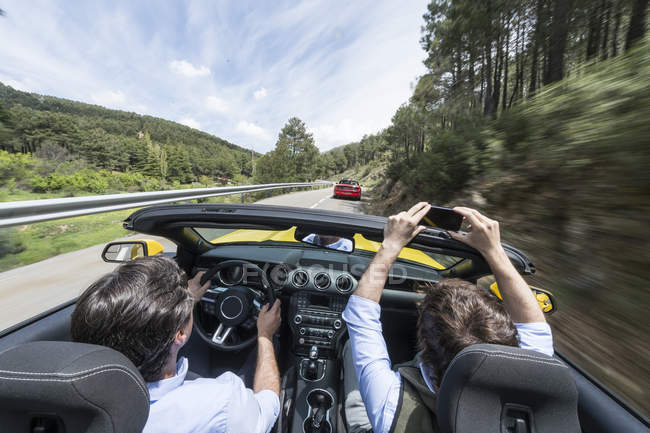 Men driving in modern fast car down road in countryside and taking photo with smartphone — Stock Photo