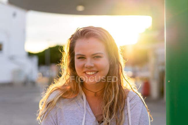 Joyeux jeune femme en tenue décontractée regardant la caméra et riant dans le dos éclairé de la lumière du coucher du soleil — Photo de stock