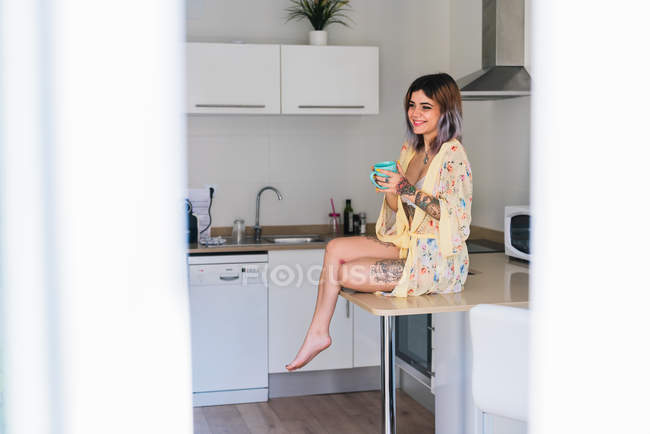 Joyeux jeune femme tatouée assise sur la table dans la cuisine avec une tasse de boisson chaude — Photo de stock