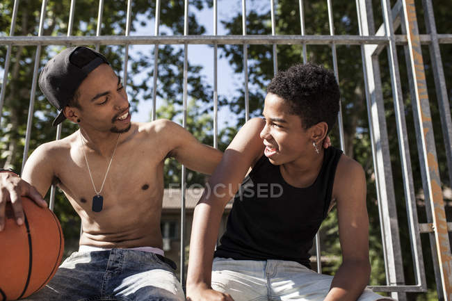 Afro jóvenes hermanos sentados con baloncesto en la cancha al aire libre y abrazando - foto de stock