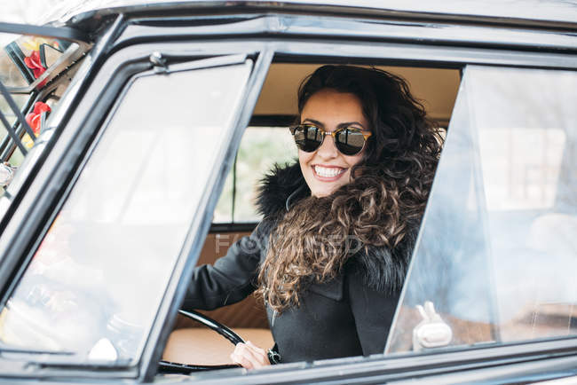 Portrait de jeune femme en manteau noir et lunettes de soleil assis à l'intérieur de la voiture — Photo de stock