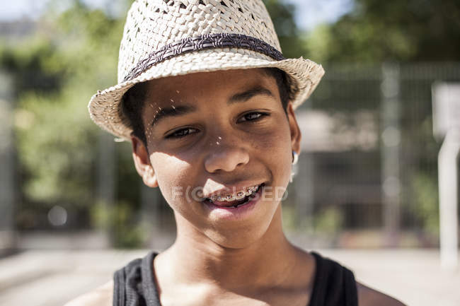 Улыбающийся мальчик в соломенной шляпе стоит на улице и смотрит в камеру — стоковое фото