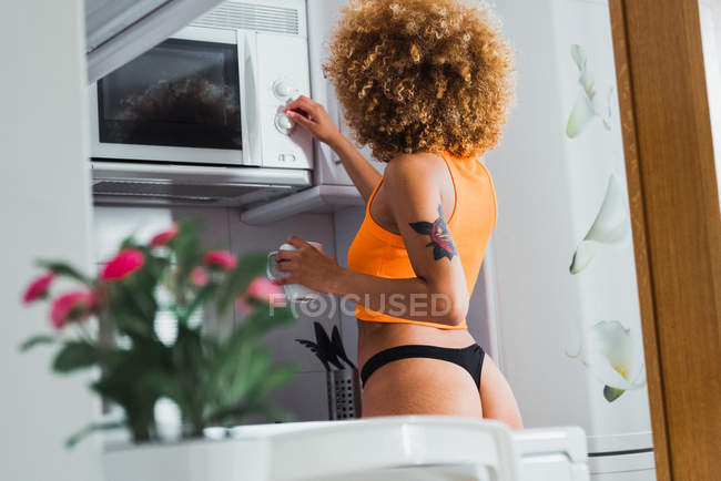 Ethnic woman in panties holding coffee cup and using microwave at home in morning — Stock Photo