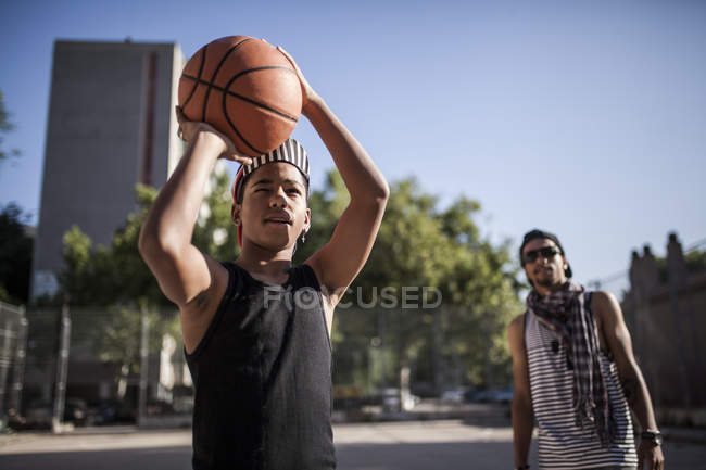 Jovem afro menino apontando basquete em cesta na quadra ao ar livre com irmão no fundo — Fotografia de Stock