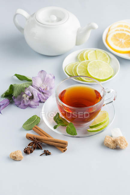 Black tea in glass cup on saucer on white background with brown sugar, cinnamon, mint, flowers and slices of citrus fruits with teapot — Stock Photo