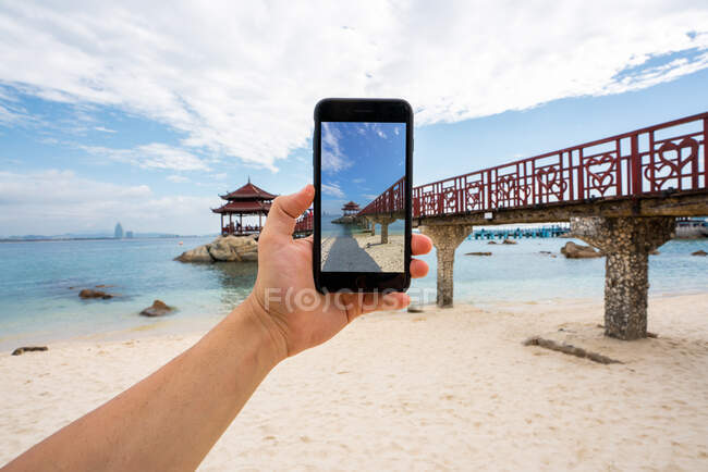 Crop hand using smartphone and taking photo of footbridge on tropical coast of Wuzhizhou Island, Hainan, China — Stock Photo