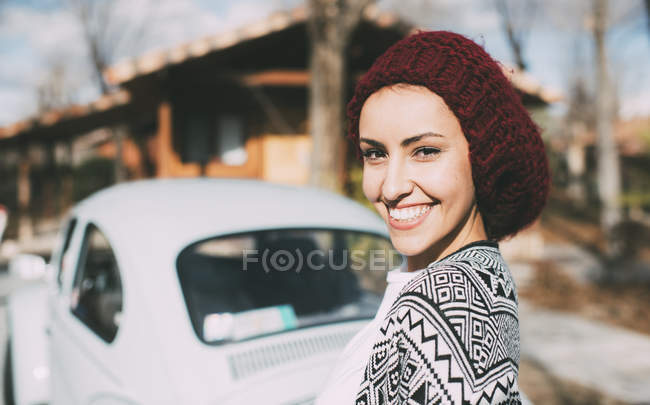 Молода жінка у вовняному капелюсі стоїть перед старою машиною і дивиться на камеру — стокове фото