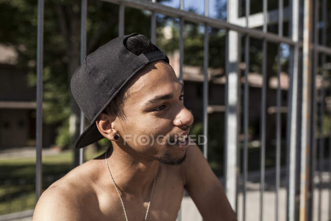 Smiling afro young boy in cap sitting against grating and looking away — Stock Photo