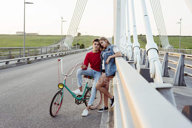 Young couple standing on bridge in countryside with vintage bicycle at sunset — Stock Photo