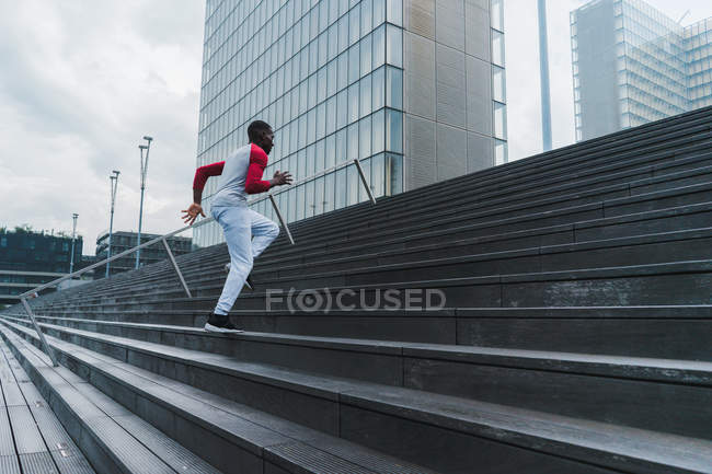 Young fit ethnic man in sportswear running up stairs with glass modern buildings on background — Stock Photo