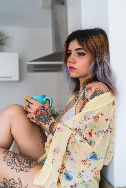 Sensuale giovane donna in abito di seta in possesso di tazza di bevanda calda e appoggiata sulla parete in cucina — Foto stock