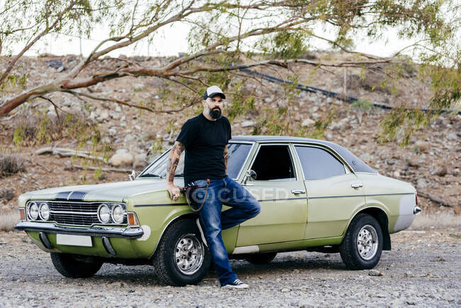 Adult handsome man leaning on green muscle car parked in nature. — Stock Photo
