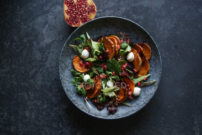 Salat mit Gemüse und Käse in Schüssel auf grauer Oberfläche mit Granatapfelhälfte — Stockfoto