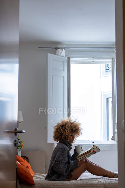 Mulher étnica relaxante com café e livro na cama em casa — Fotografia de Stock
