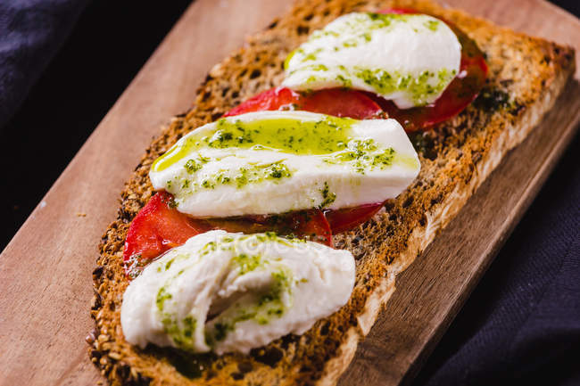 Крупный план тоста из золотого хлеба с помидорами и белым сыром, увенчанным зеленым соусом на деревянной доске — стоковое фото