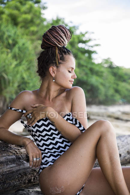 Dreamy woman in patterned swimsuit sitting on roots on beach — Stock Photo