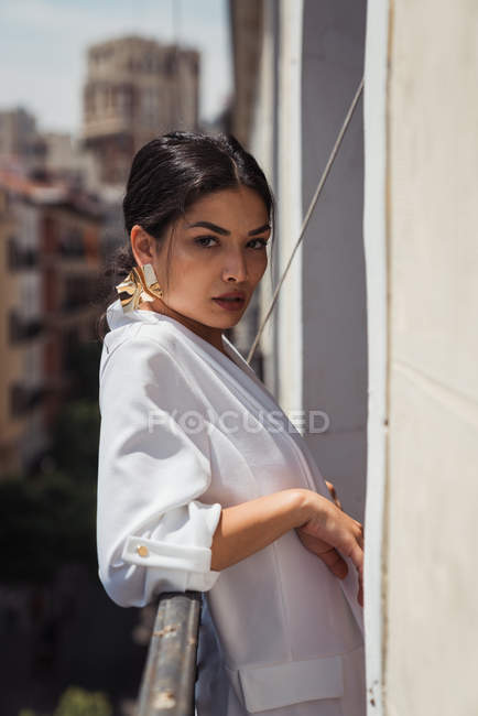 Sensuale donna bruna in elegante giacca bianca e orecchini dorati guardando la fotocamera mentre si appoggia sulla recinzione del balcone — Foto stock