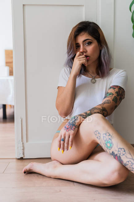 Sinnlich tätowierte junge Frau sitzt auf dem Boden und schaut zu Hause in die Kamera — Stockfoto