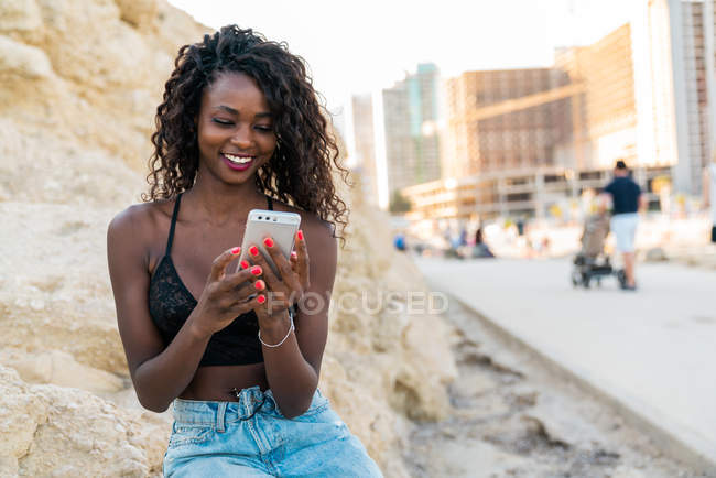 Cheerful black woman using phone while sitting on rock outside — Stock Photo